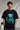 Man wearing a black BROKEN CODE oversized t-shirt with a glitch art graphic of eyes and text. Features a chain necklace and dark wash jeans. Model is posed against a studio backdrop.