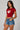 Model wearing a red CROPTOP Cherry crop top with a cute cherry and bow graphic, paired with denim shorts. The top has a flattering cropped fit.