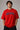 Male model wearing a red oversized t-shirt with 'Believe' printed in navy blue and white collegiate-style lettering.