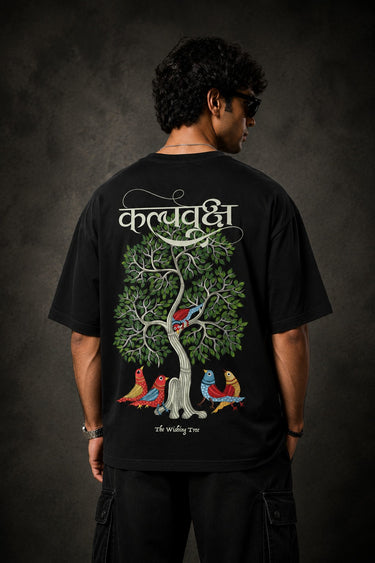 Man wearing a black Kalpvruksh oversized t-shirt with a colorful tree and bird graphic print on the back. Features Hindi script and 'The Wishing Tree' text. Streetwear style.