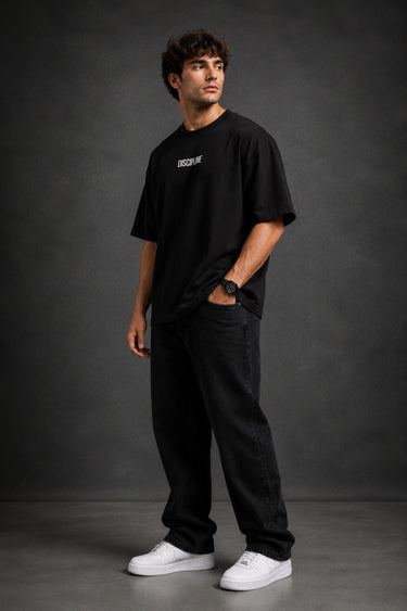 Man wearing a black DISCIPLINE WILD WOLF oversized t-shirt with white 'DISCIPLINE' text, dark wash jeans, and white sneakers, sitting on stairs.