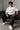 Man wearing white F1 Official oversized t-shirt with red F1 logo, black wide-leg jeans, and white sneakers, sitting on a black leather couch.
