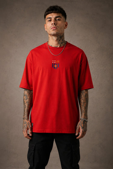 Man wearing a red FCB Messi fan oversized t-shirt, black pants, and white sneakers, sitting on a brown couch. - red