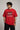 Man wearing a red PROJECTOR HANUMANJI oversized t-shirt with black pants and white sneakers, sitting on metal stairs.