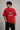 Man wearing a red PROJECTOR HANUMANJI oversized t-shirt with black pants and white sneakers, sitting on metal stairs.
