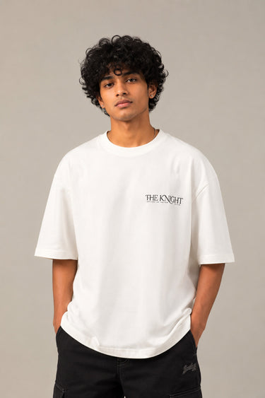 Man wearing a white 'THE KNIGHT' oversized t-shirt and black pants, sitting on concrete stairs. Features short curly hair and white sneakers. - white