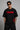 Man wearing a black oversized t-shirt with a red 'SPIDERMAN' puff print graphic and a red spider logo on the side. Model is wearing sunglasses and cargo pants.