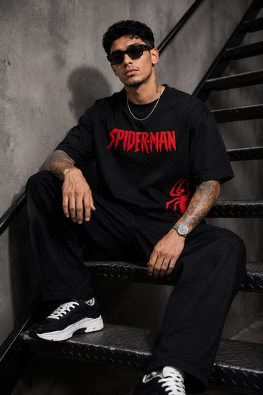 Man wearing a black oversized t-shirt with a red 'SPIDERMAN' puff print graphic and a red spider logo on the side. Model is wearing sunglasses and cargo pants.