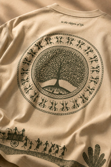 Close-up of a Rooted oversized t-shirt in beige, featuring a dark green circular graphic of a tree of life with tribal figures, and text 'in the rhfym of life'.