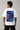 Man wearing a white VIRAT MCG 18 Vs PAK oversized t-shirt with a blue galaxy-themed '18' graphic. He is sitting on steps, showcasing the relaxed fit and streetwear style. - white