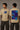 Man wearing a white VIRAT MCG 18 Vs PAK oversized t-shirt with a blue galaxy-themed '18' graphic. He is sitting on steps, showcasing the relaxed fit and streetwear style. - beige