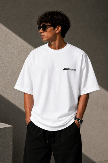 Man wearing a white oversized BMW M "530i E60" graphic t-shirt and black baggy jeans, standing against a textured concrete wall.