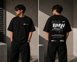Man wearing a white oversized BMW M "530i E60" graphic t-shirt and black baggy jeans, standing against a textured concrete wall. - black
