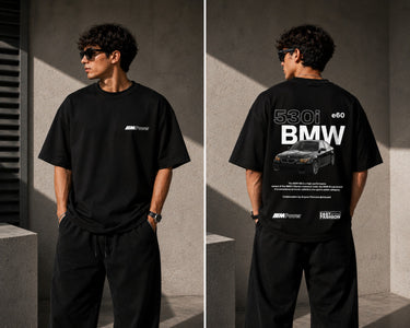 Man wearing a white oversized BMW M "530i E60" graphic t-shirt and black baggy jeans, standing against a textured concrete wall. - black