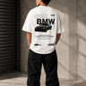 Man wearing a white oversized BMW M "530i E60" graphic t-shirt and black baggy jeans, standing against a textured concrete wall.