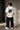 Man wearing a white oversized BMW M "530i E60" graphic t-shirt and black baggy jeans, standing against a textured concrete wall.