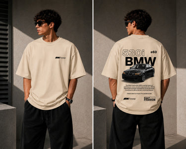 Man wearing a white oversized BMW M "530i E60" graphic t-shirt and black baggy jeans, standing against a textured concrete wall. - beige