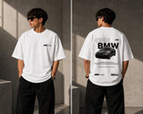 Man wearing a white oversized BMW M "530i E60" graphic t-shirt and black baggy jeans, standing against a textured concrete wall. - white