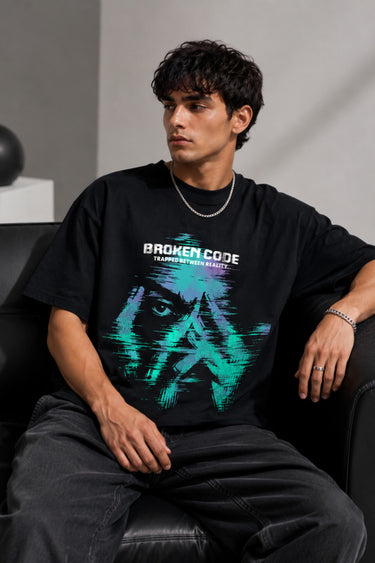 Man wearing a black BROKEN CODE oversized t-shirt with a glitch art graphic of eyes and text. Features a chain necklace and dark wash jeans. Model is posed against a studio backdrop.