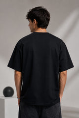 Man wearing a black BROKEN CODE oversized t-shirt with a glitch art graphic of eyes and text. Features a chain necklace and dark wash jeans. Model is posed against a studio backdrop.