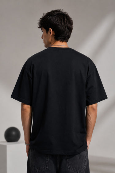 Man wearing a black BROKEN CODE oversized t-shirt with a glitch art graphic of eyes and text. Features a chain necklace and dark wash jeans. Model is posed against a studio backdrop.