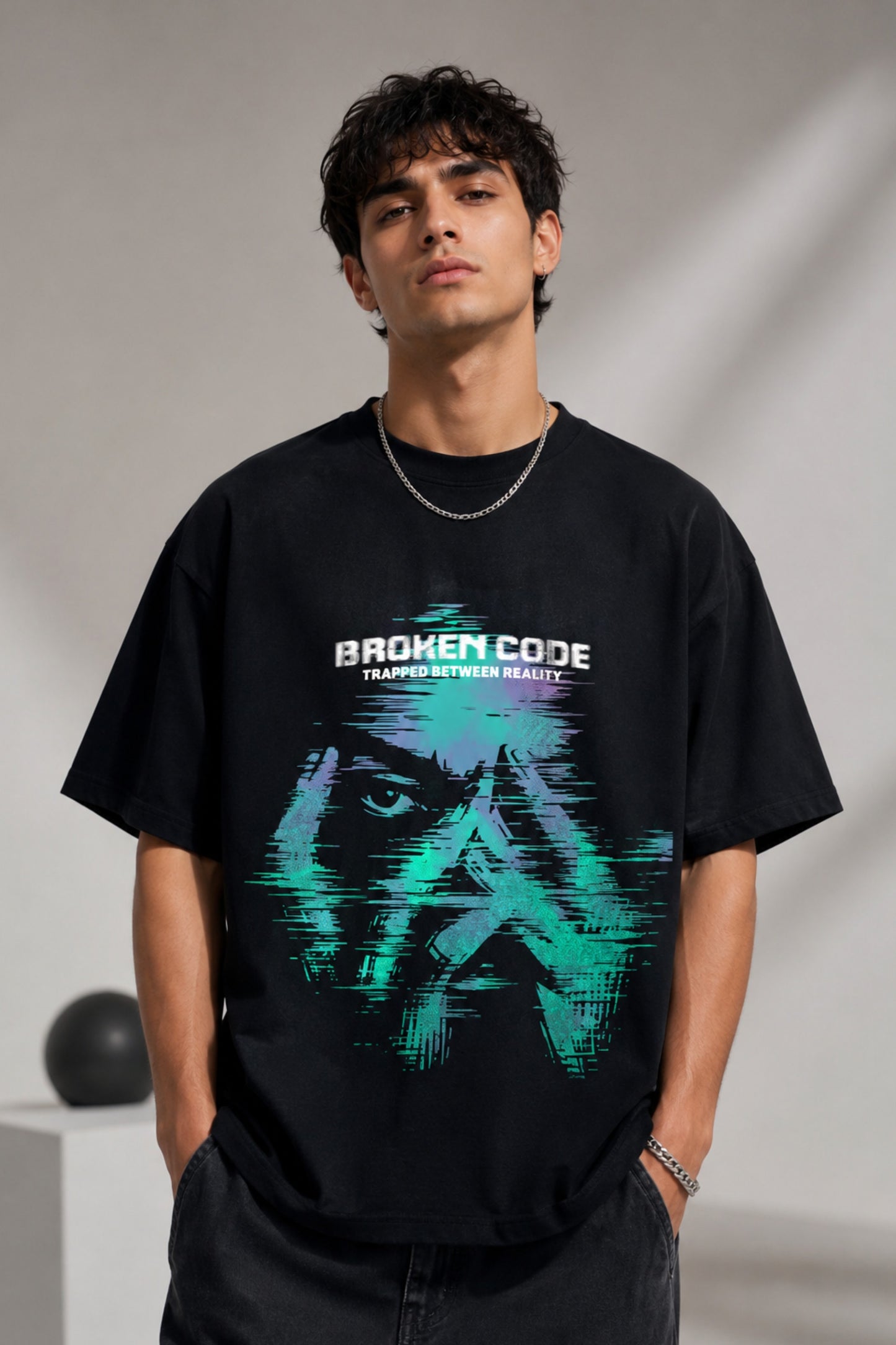 Man wearing a black BROKEN CODE oversized t-shirt with a glitch art graphic of eyes and text. Features a chain necklace and dark wash jeans. Model is posed against a studio backdrop.
