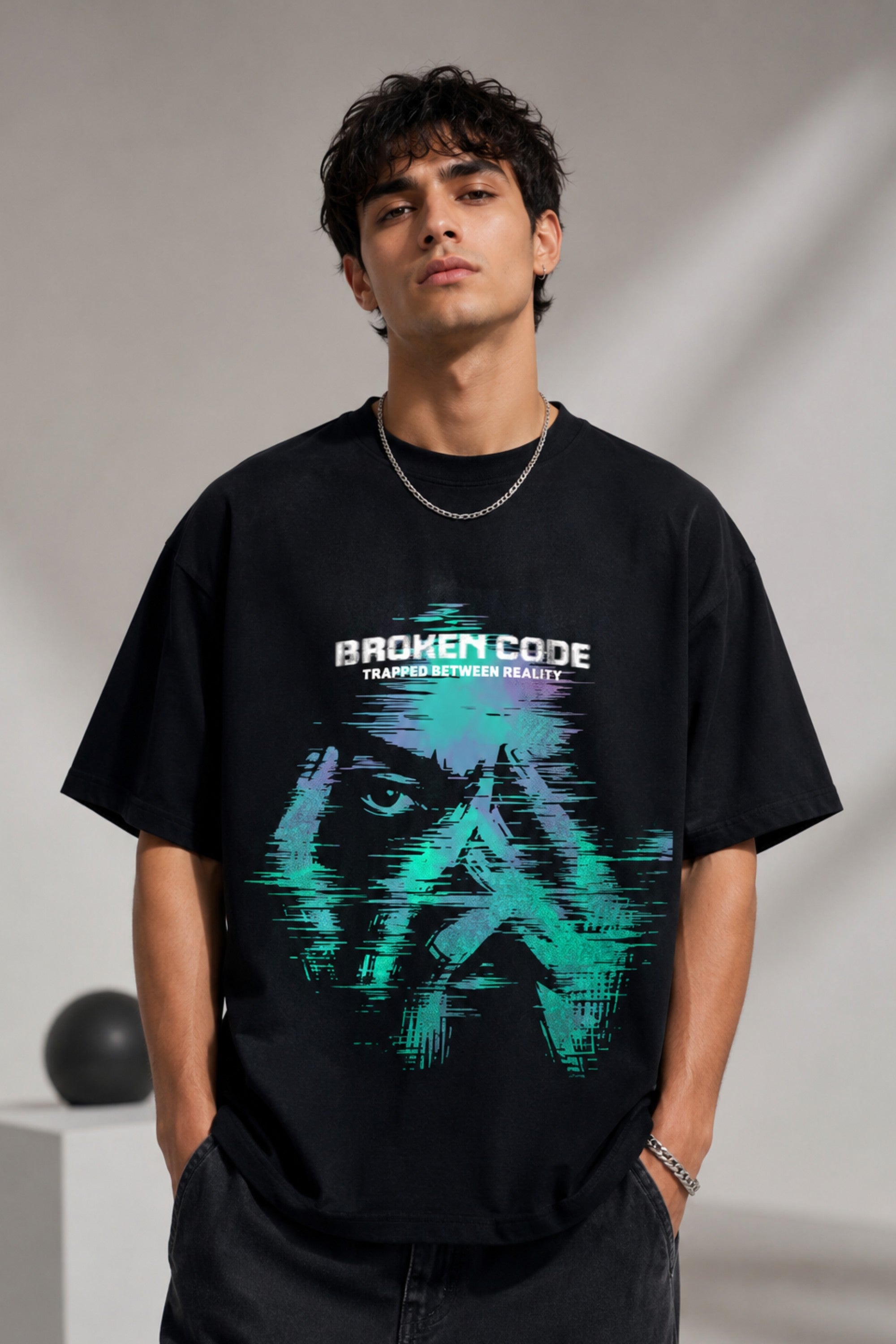 Man wearing a black BROKEN CODE oversized t-shirt with a glitch art graphic of eyes and text. Features a chain necklace and dark wash jeans. Model is posed against a studio backdrop.