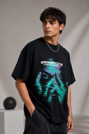 Man wearing a black BROKEN CODE oversized t-shirt with a glitch art graphic of eyes and text. Features a chain necklace and dark wash jeans. Model is posed against a studio backdrop.