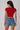 Model wearing a red CROPTOP Cherry crop top with a cute cherry and bow graphic, paired with denim shorts. The top has a flattering cropped fit.