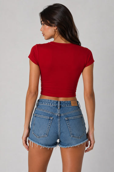 Model wearing a red CROPTOP Cherry crop top with a cute cherry and bow graphic, paired with denim shorts. The top has a flattering cropped fit.