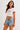 Woman wearing a white crop top with red trim and "New York" graphic, paired with denim shorts, sitting on a textured armchair.
