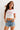 Woman wearing a white crop top with red trim and "New York" graphic, paired with denim shorts, sitting on a textured armchair.