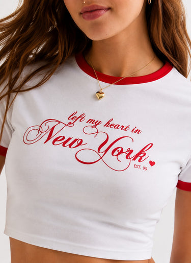 Woman in white ringer crop top with red trim and 'New York' graphic, paired with denim shorts, sitting on a couch.
