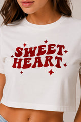 Model wearing a white "Sweet Heart" graphic oversized croptop t-shirt with red distressed lettering and star accents. Streetwear fashion.