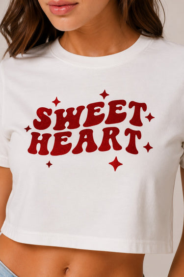 Model wearing a white "Sweet Heart" graphic oversized croptop t-shirt with red distressed lettering and star accents. Streetwear fashion.