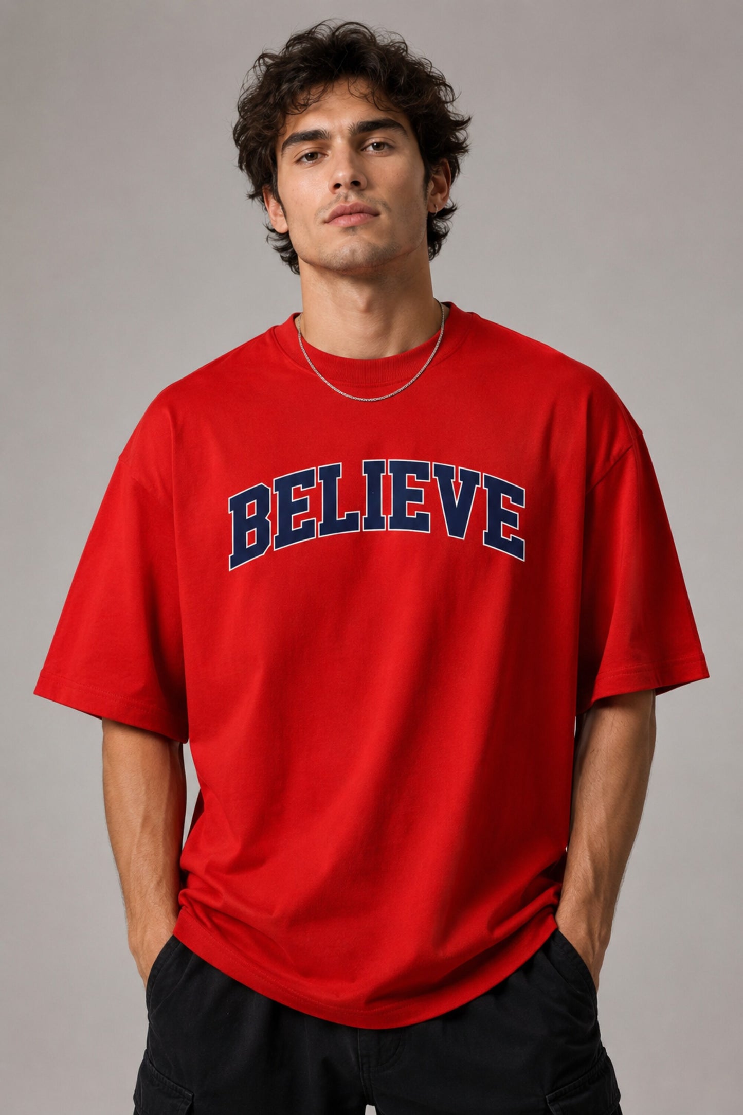 Male model wearing a red oversized t-shirt with 'Believe' printed in navy blue and white collegiate-style lettering.