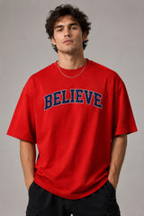 Male model wearing a red oversized t-shirt with 'Believe' printed in navy blue and white collegiate-style lettering.