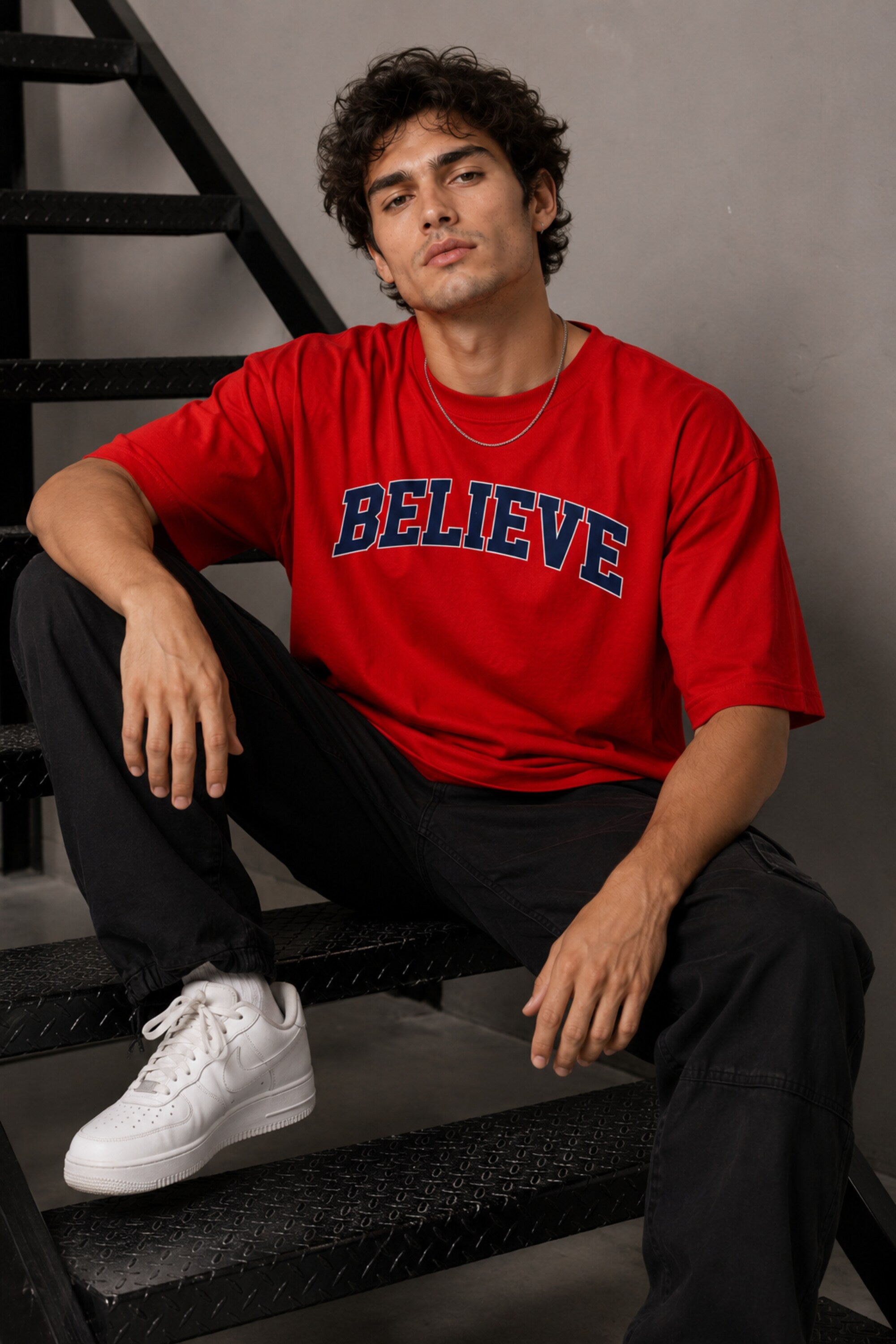 Male model wearing a red oversized t-shirt with 'Believe' printed in navy blue and white collegiate-style lettering.