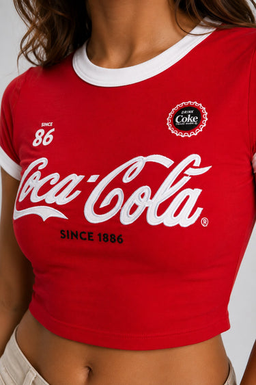 Back view of a woman wearing a red Coco-Cola crop top with white trim and beige high-waisted pants against a white background.