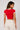 Back view of a woman wearing a red Coco-Cola crop top with white trim and beige high-waisted pants against a white background.