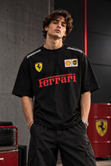 A man wearing a black Ferrari crewneck t-shirt with red and yellow sponsor logos, standing in a garage-like setting with red barrels in the background.