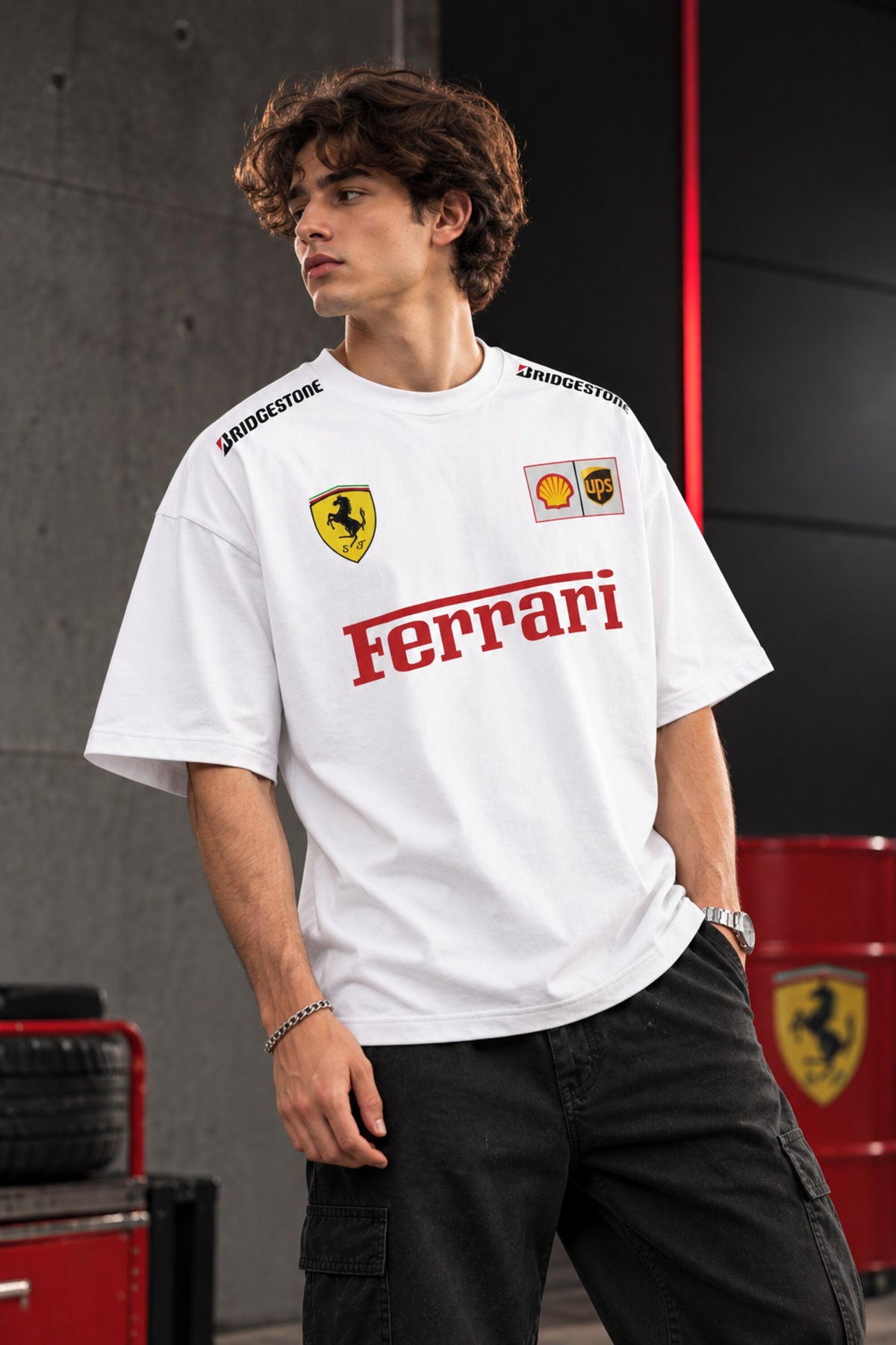 A man wearing a white Ferrari crewneck t-shirt with red and yellow sponsor logos, paired with black cargo pants, standing in a garage-like setting with red Ferrari-branded equipment.