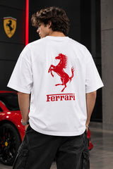 A man wearing a white t-shirt with a large red Ferrari prancing horse logo and the word 'Ferrari' printed on the back, standing in a garage-like setting with a red car visible on the left.