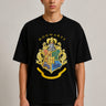 Black oversized t-shirt with the Hogwarts crest and 'Hogwarts' text in yellow, worn by a person. Features detailed coat of arms with lions, snakes, badgers, and eagles.