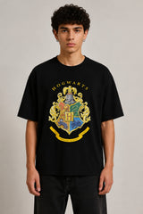 Black oversized t-shirt with the Hogwarts crest and 'Hogwarts' text in yellow, worn by a person. Features detailed coat of arms with lions, snakes, badgers, and eagles.