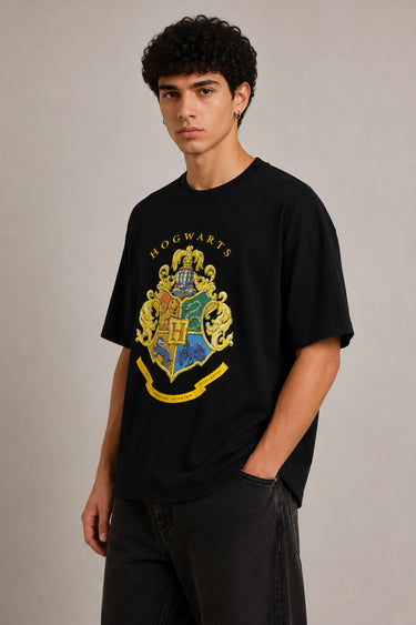 Black oversized t-shirt with the Hogwarts crest and 'Hogwarts' text in yellow, worn by a person. Features detailed coat of arms with lions, snakes, badgers, and eagles.