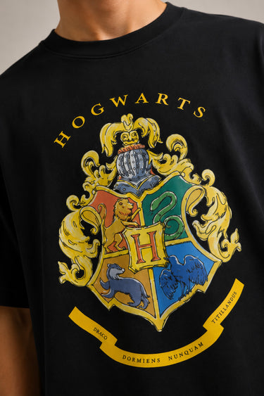 Black oversized t-shirt with the Hogwarts crest and 'Hogwarts' text in yellow, worn by a person. Features detailed coat of arms with lions, snakes, badgers, and eagles.