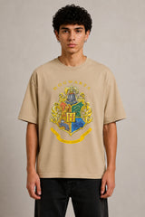 Black oversized t-shirt with the Hogwarts crest and 'Hogwarts' text in yellow, worn by a person. Features detailed coat of arms with lions, snakes, badgers, and eagles. - beige