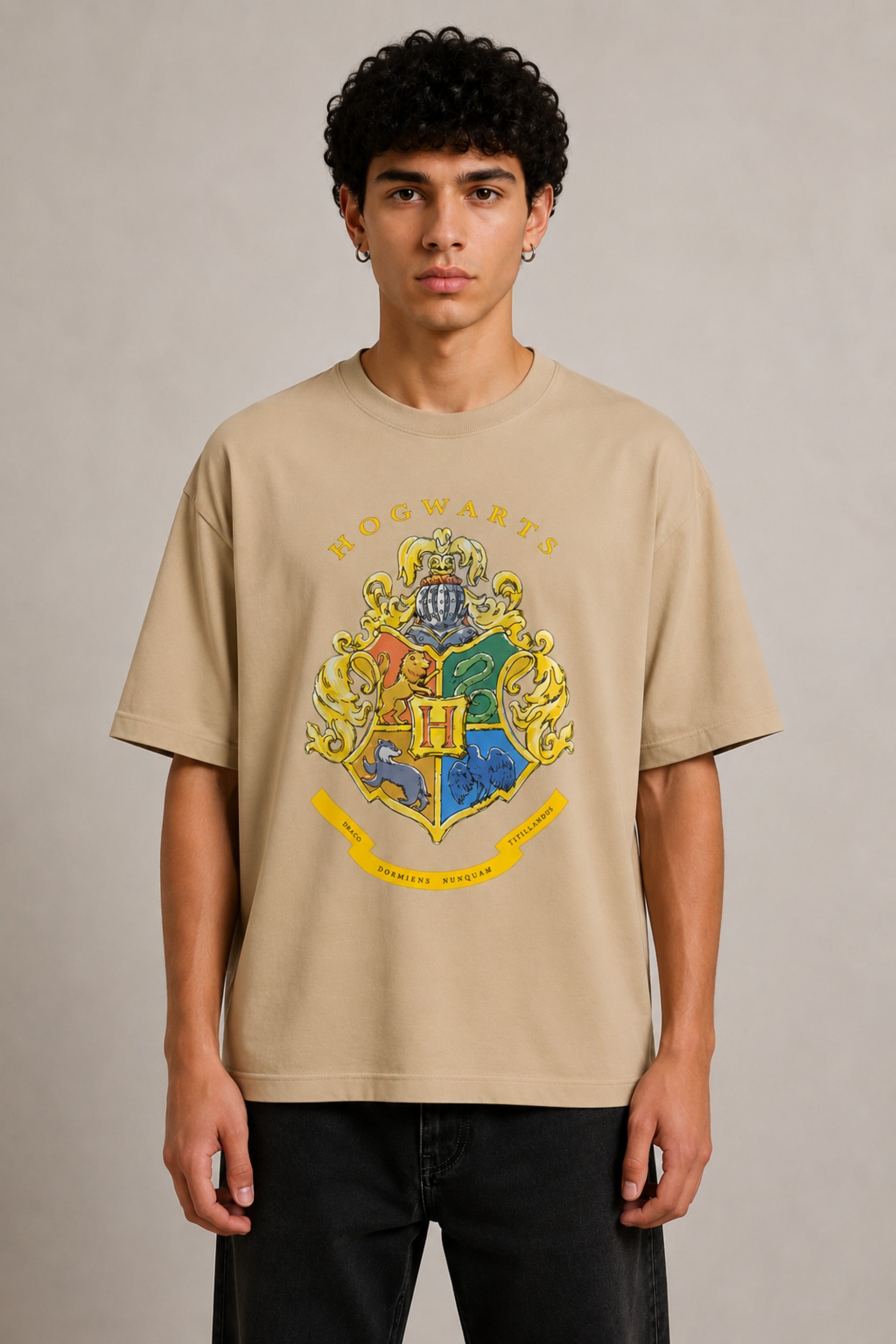 Black oversized t-shirt with the Hogwarts crest and 'Hogwarts' text in yellow, worn by a person. Features detailed coat of arms with lions, snakes, badgers, and eagles. - beige