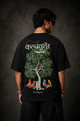 Man wearing a black Kalpvruksh oversized t-shirt with a colorful tree and bird graphic print on the back. Features Hindi script and 'The Wishing Tree' text. Streetwear style.